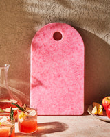 Pink recycled chopping board with marbled pattern, arched top, surrounded by apples and drinks